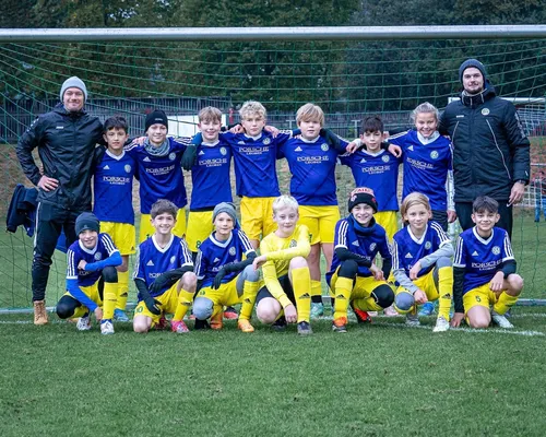 Eine Jugendfußballmannschaft in blauen und gelben Trikots posiert mit zwei Trainern auf dem Spielfeld für ein Foto.