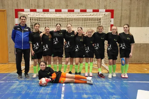 Eine Gruppe junger Mädchen in identischen Trikots steht hinter einem Tornetz. Sie scheinen für ein Foto zu posieren. Das Mädchen vorne liegt auf dem Boden und hält einen Fußball.