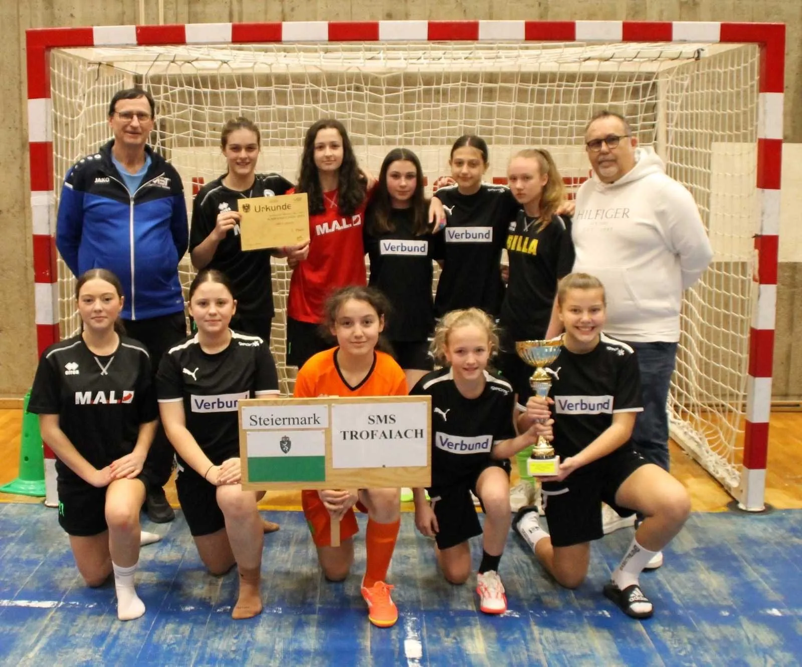 Eine Sportmannschaft, wahrscheinlich junge Mädchen, posiert für ein Foto mit Trophäen und Urkunden vor einem Tor. Sie tragen passende schwarze Trikots mit weißen Texten. Dahinter stehen zwei Erwachsene, einer hält eine Urkunde.