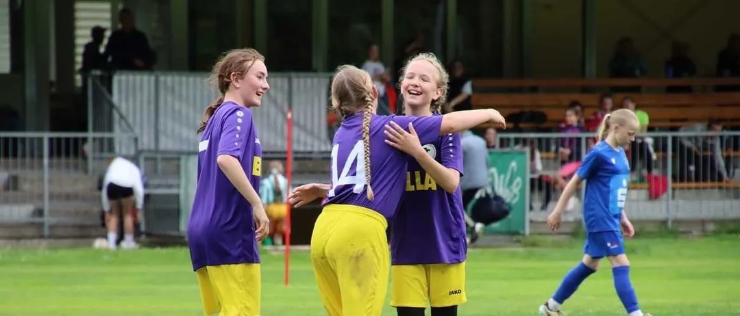 Drei Mädchen in lila und gelben Trikots stehen auf einem Fußballfeld, lächeln und umarmen sich. Hinter ihnen sitzen Zuschauer auf Tribünen.
