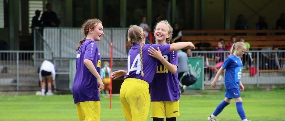 Drei Mädchen in lila und gelben Trikots stehen auf einem Fußballfeld, lächeln und umarmen sich. Hinter ihnen sitzen Zuschauer auf Tribünen.