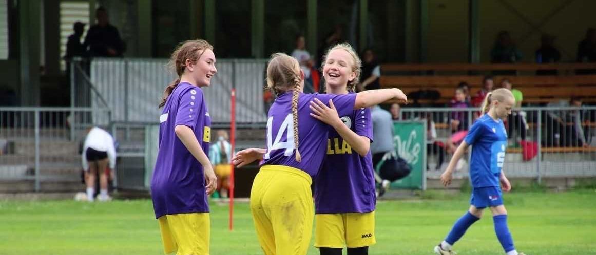 Drei Mädchen in lila und gelben Trikots stehen auf einem Fußballfeld, lächeln und umarmen sich. Hinter ihnen sitzen Zuschauer auf Tribünen.