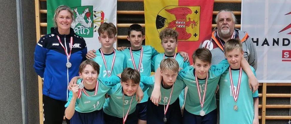 Eine Jugendhandballmannschaft mit Medaillen posiert für ein Foto. Einige sitzen und andere stehen, alle tragen blaue Uniformen und Medaillen. Zwei Erwachsene stehen hinter ihnen, einer hält ein Zertifikat.