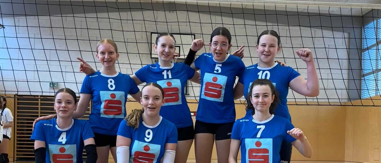 Eine Gruppe von weiblichen Volleyballspielerinnen in blauen Uniformen posiert für ein Bild in einer Turnhalle. Sie halten einen Volleyball.
