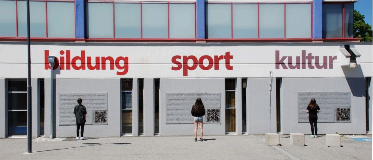 Zwei Personen stehen vor einem Gebäude, auf dem 'lung sport ku' steht. Eine hält ein Smartphone und die andere steht neben einem Pfosten.