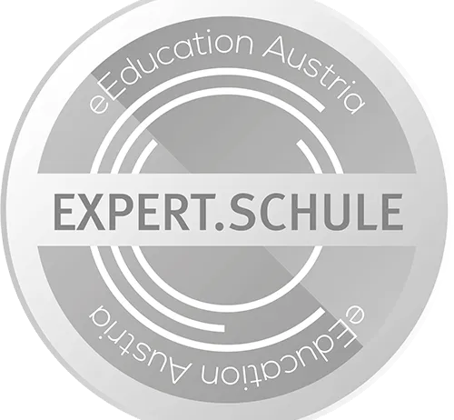 Ein kreisförmiges Abzeichen mit grauem Hintergrund und weißem Rand. Oben steht 'eEducation Austria' und in der Mitte 'EXPERT.SCHULE', mit einem geschwungenen weißen Liniendesign.