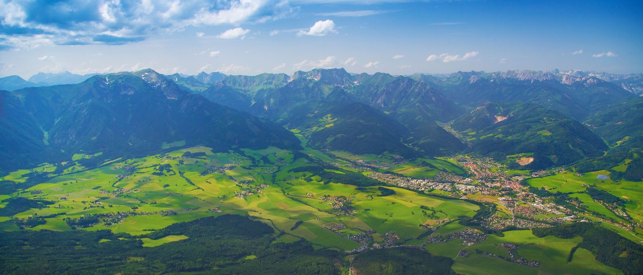 Luftaufnahme eines grünen Tals mit grünen Bergen und einer Stadt darunter, unter einem blauen Himmel mit Wolken.