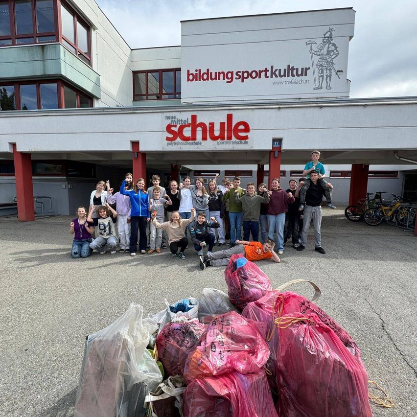 Schüler posieren vor einem Gebäude namens 'Bildung Sport Kultur Schule' mit Säcken gesammelten Mülls davor.