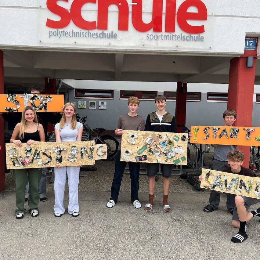 Eine Gruppe von Schülern steht vor einem Gebäude mit Schildern, die 'STOP', 'WASTEING', 'START' und 'CANVAS' sagen. Sie halten ihre Schilder hoch, lächeln und posieren für ein Foto. Dahinter befindet sich ein Gebäude mit dem Schild 'SCHULE'.