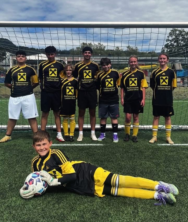 Eine Jugendfußballmannschaft posiert für ein Foto mit einem Torwart, der kniet und einen Fußball vor einem Tor hält.