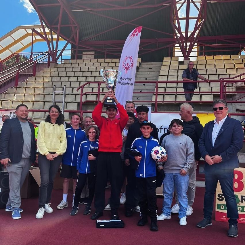 Eine Gruppe von Menschen steht in einem Stadion und hält eine Trophäe. Ein Mann in einem roten Jackett hält die Trophäe. Ein Junge mit einem Fußball steht vor ihm. Leere Tribünen sind hinter ihnen.