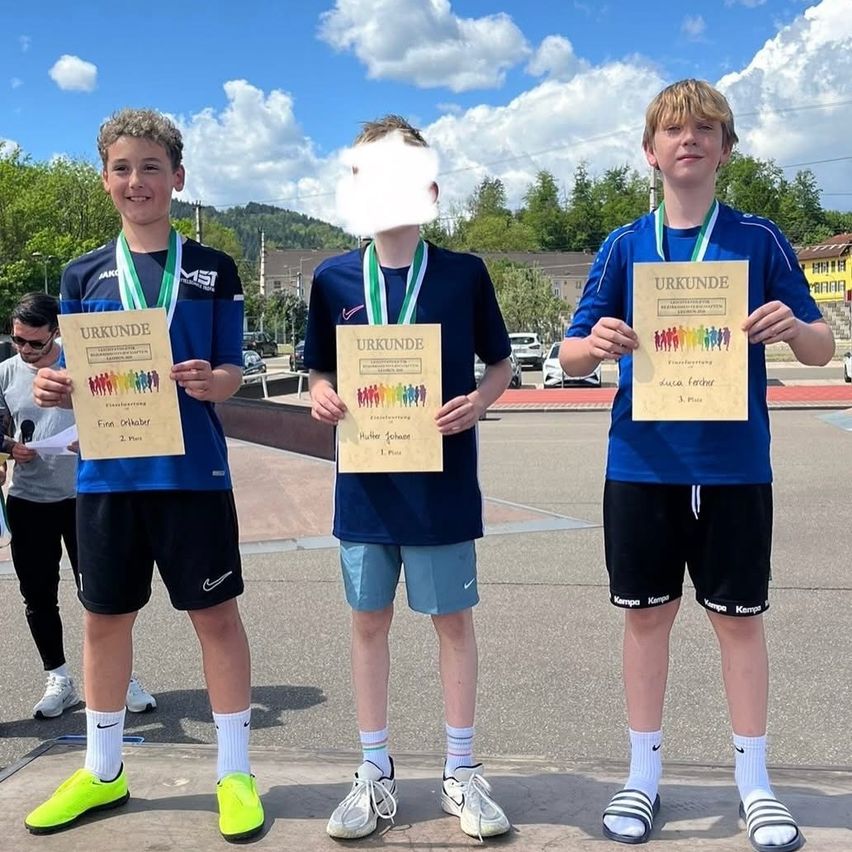 Drei Jungen mit Medaillen um den Hals stehen in einem Außenbereich. Sie halten Urkunden mit einem Bild von Menschen darauf. Der Junge auf der rechten Seite hat ein verschwommenes Gesicht.