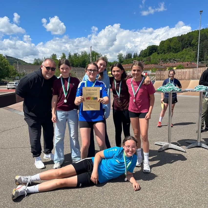 Eine Gruppe junger Athletinnen, darunter ein Mädchen mit einem Zertifikat, posiert für ein Foto mit Medaillen um den Hals an einem sonnigen Tag.