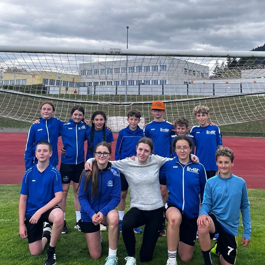 Eine Gruppe junger Fußballspieler in blauen Uniformen posiert für ein Foto auf einem Rasenfeld mit einem Fußballtor im Hintergrund.