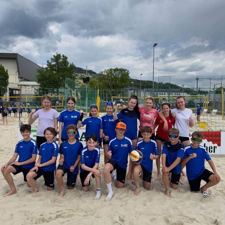 Eine Beachvolleyballmannschaft in blauen und weißen Uniformen posiert für ein Gruppenfoto auf dem Sand. Dahinter ist ein Volleyballnetz aufgebaut, mit stehengebliebenen Personen und Bäumen im Hintergrund.