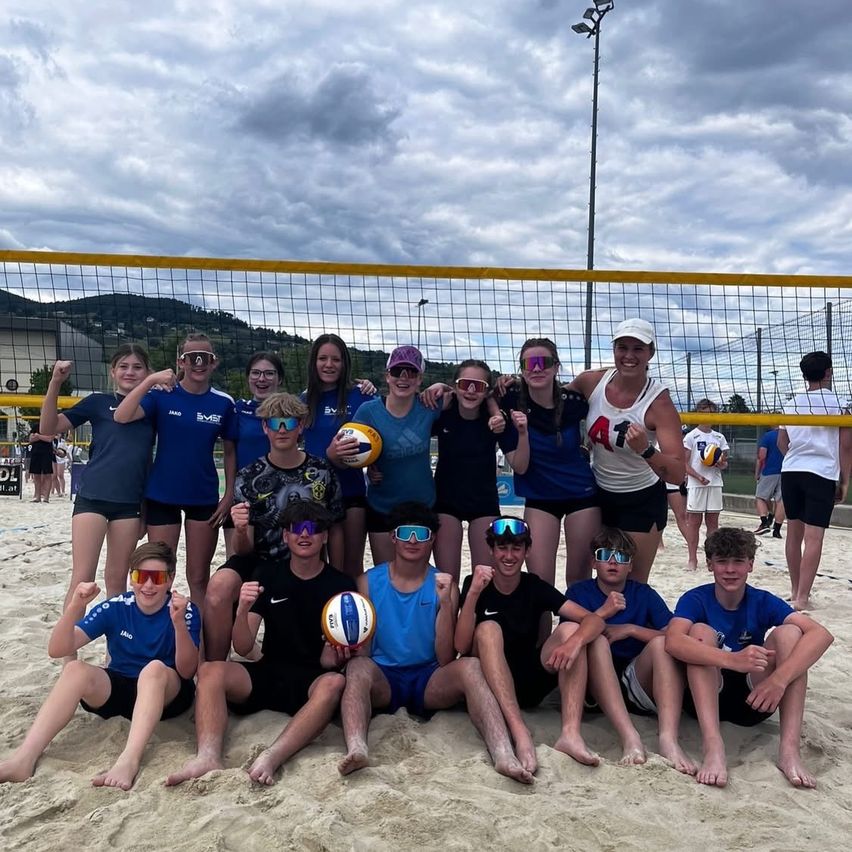 Eine Gruppe von Menschen in Sonnenbrillen und Sportkleidung posiert für ein Foto am Strand mit einem Volleyballnetz im Hintergrund.