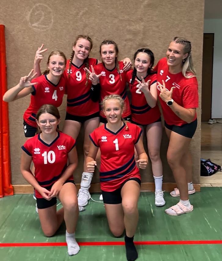 Eine Gruppe junger Frauen in roten Volleyballtrikots posiert für ein Foto. Sie lächeln und machen Friedenszeichen. Sie stehen auf einem grünen Boden.