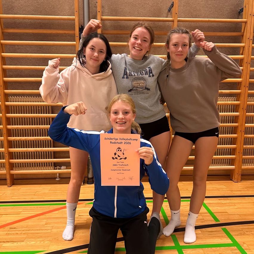 Vier Mädchen in Sportkleidung posieren für ein Foto in einer Turnhalle. Eine hält ein Zertifikat, während die anderen hinter ihr anfeuern. Die Holzwand und die Heizkörper im Hintergrund deuten auf eine Sporteinrichtung hin.