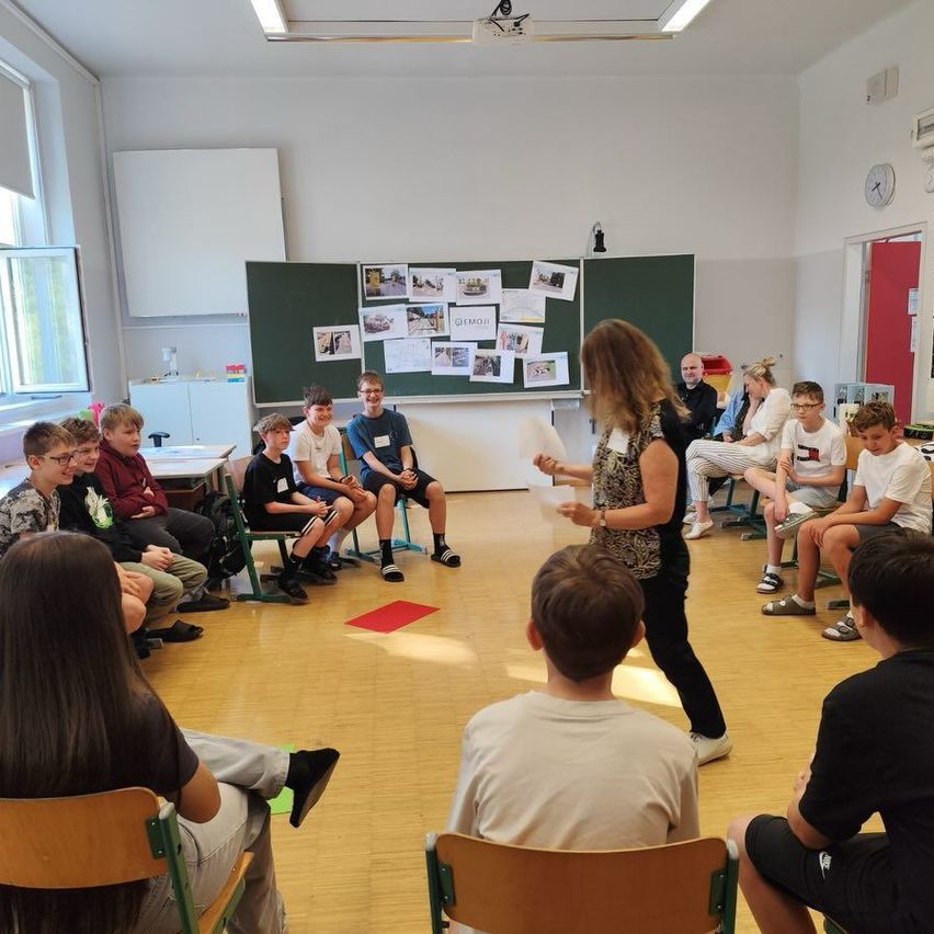 Eine Frau steht in der Mitte eines Klassenzimmers, in dem Schüler auf Stühlen sitzen. Das Klassenzimmer hat eine Bulletin-Board und eine Uhr an der Wand. Die Frau hält ein Papier in der Hand.