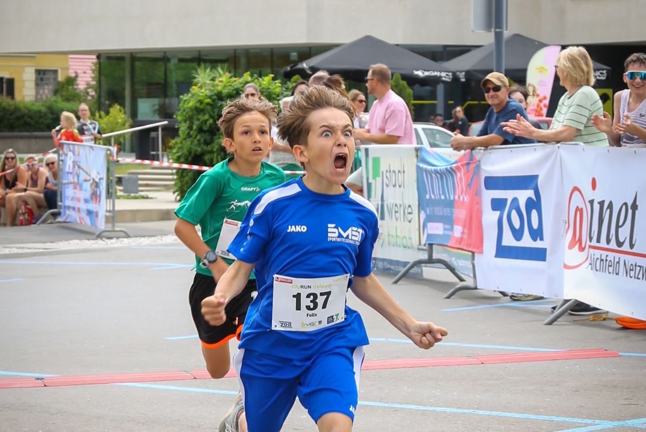 Zwei junge Jungen nehmen an einem Rennen teil, einer trägt ein blaues Trikot mit der Nummer 137 und der andere ist grün. Zuschauer beobachten hinter einem Zaun.