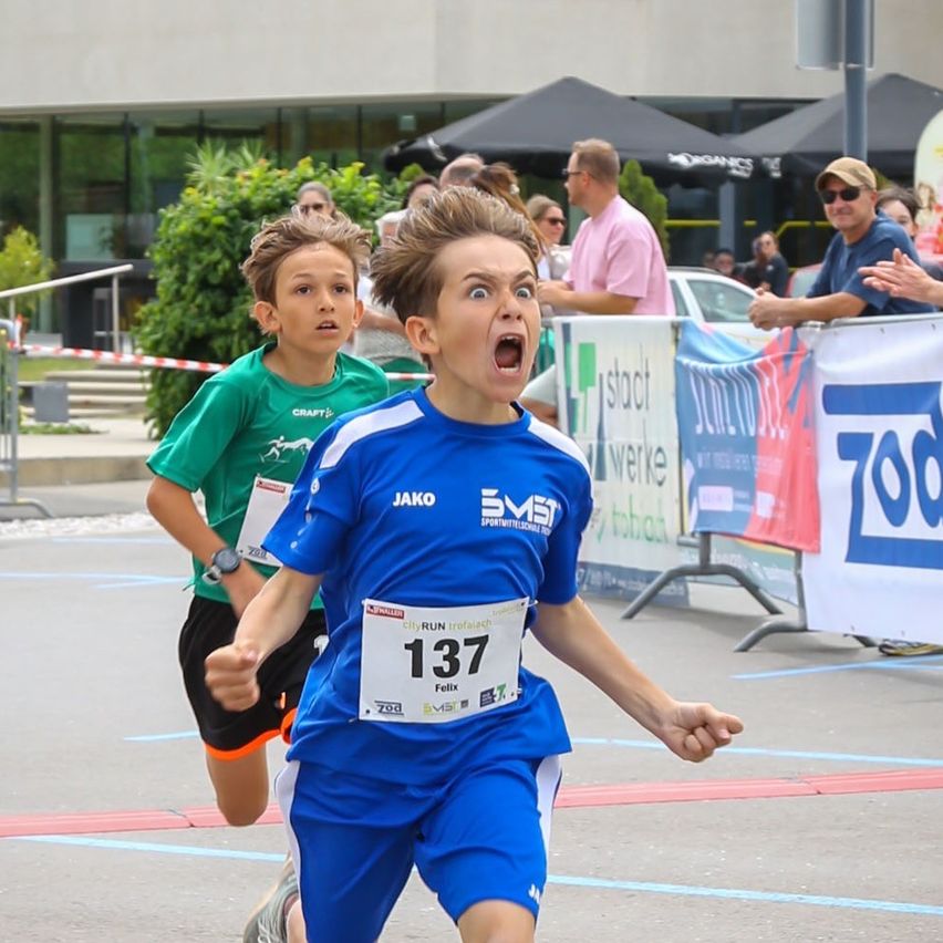 Zwei junge Jungen nehmen an einem Rennen teil, einer trägt ein blaues Trikot mit der Nummer 137 und der andere ist grün. Zuschauer beobachten hinter einem Zaun.