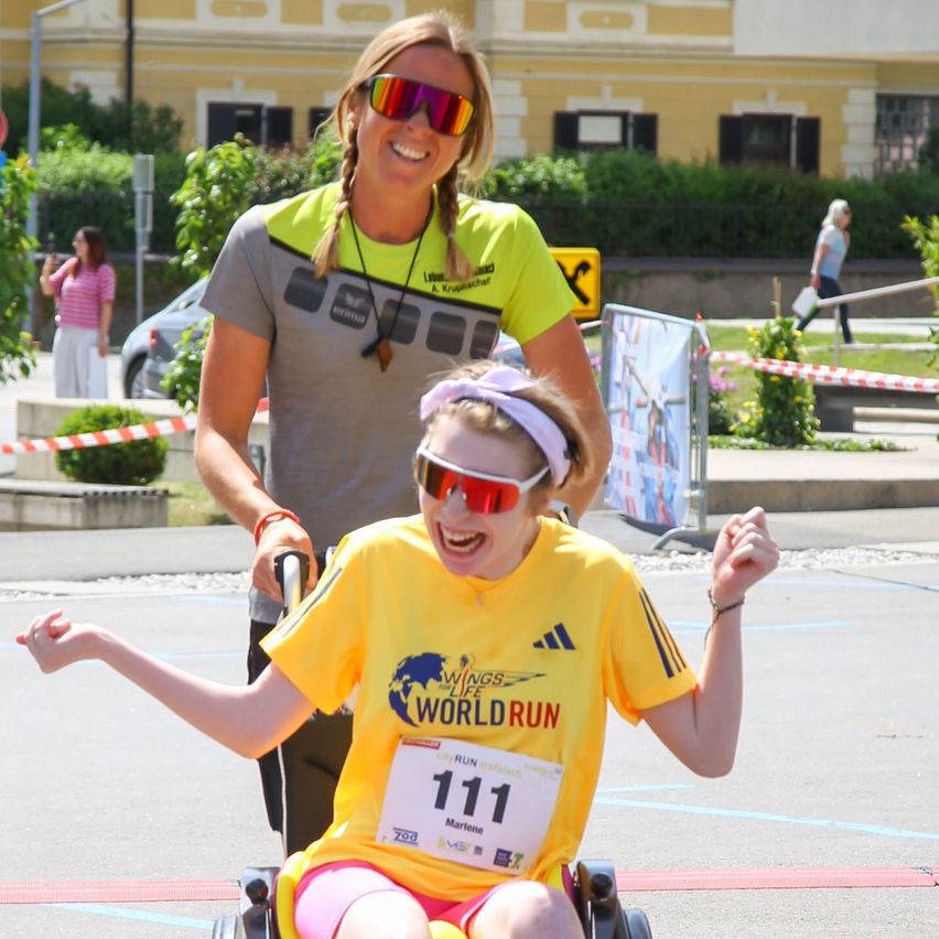 Ein Mädchen im Rollstuhl, das ein gelbes Shirt mit dem Wings for Life World Run-Logo trägt, lächelt. Hinter ihr schiebt eine Frau sie und lächelt ebenfalls. Sie befinden sich auf einer Straße, mit einem gelben Gebäude und Grünpflanzen im Hintergrund.