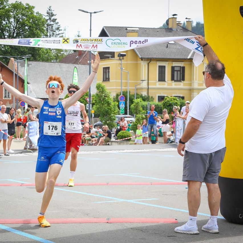 Ein Läufer mit Startnummer 176 überquert die Ziellinie eines Marathons, während ein Mann ein Band hält. Zuschauer beobachten im Hintergrund.