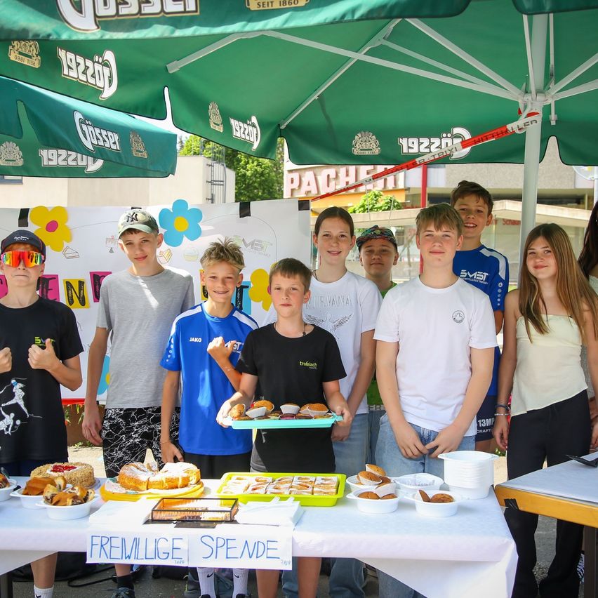 Eine Gruppe von Kindern und Jugendlichen steht hinter einem Tisch mit Essen. Im Hintergrund befinden sich Schirme mit Text darauf.