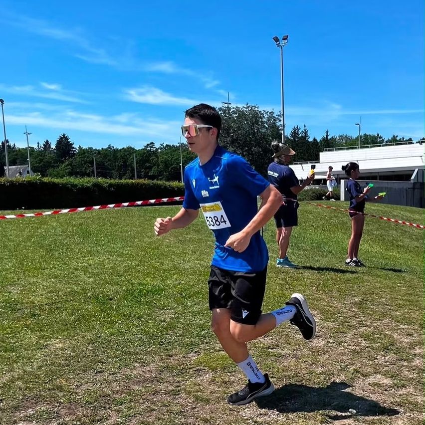 Ein Läufer mit blauem Shirt und Sonnenbrille läuft ein Rennen auf einem Grasfeld. Im Hintergrund stehen zwei Personen und halten etwas. Bäume und Straßenlaternen sind in der Ferne.