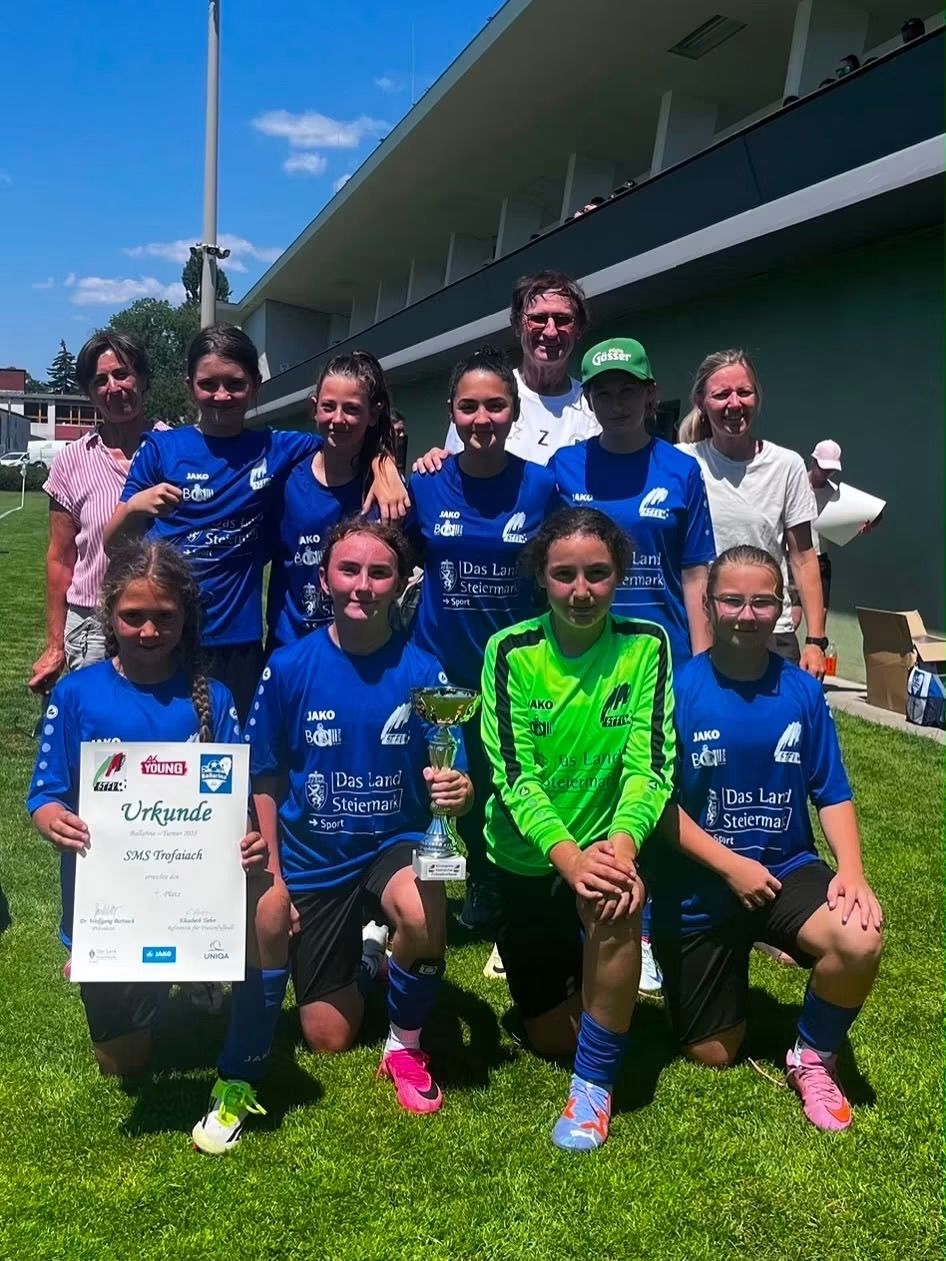 Ein Mädchenfußballteam in blauen Trikots, darunter eines in grün, posiert mit einem Pokal auf einem Rasenfeld, mit einem Gebäude und Bäumen im Hintergrund.