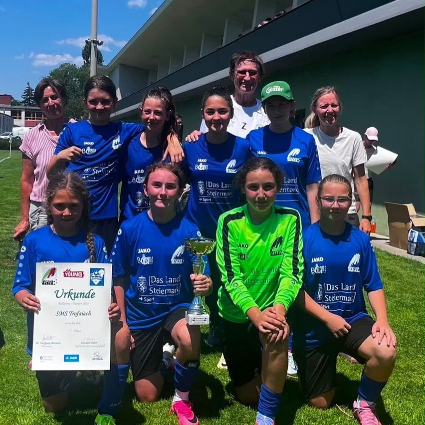 Ein Mädchenfußballteam in blauen Trikots, darunter eines in grün, posiert mit einem Pokal auf einem Rasenfeld, mit einem Gebäude und Bäumen im Hintergrund.
