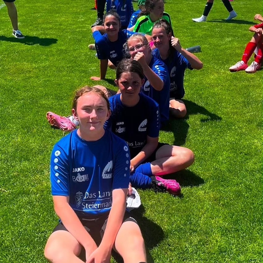 Eine Gruppe junger Mädchen in blauen Trikots sitzt auf einem grünen Rasen. Sie scheinen sich nach einem Fußballspiel auszuruhen.