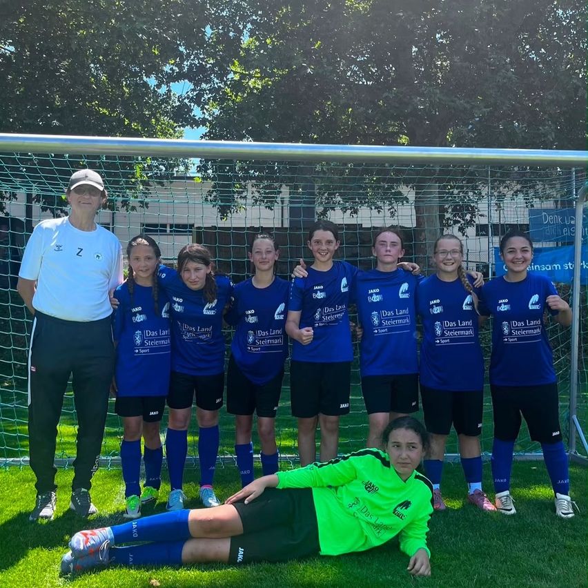 Ein Fußballteam junger Mädchen und ihr Trainer, posieren für ein Foto auf einem Rasenfeld mit einem Tor im Hintergrund.