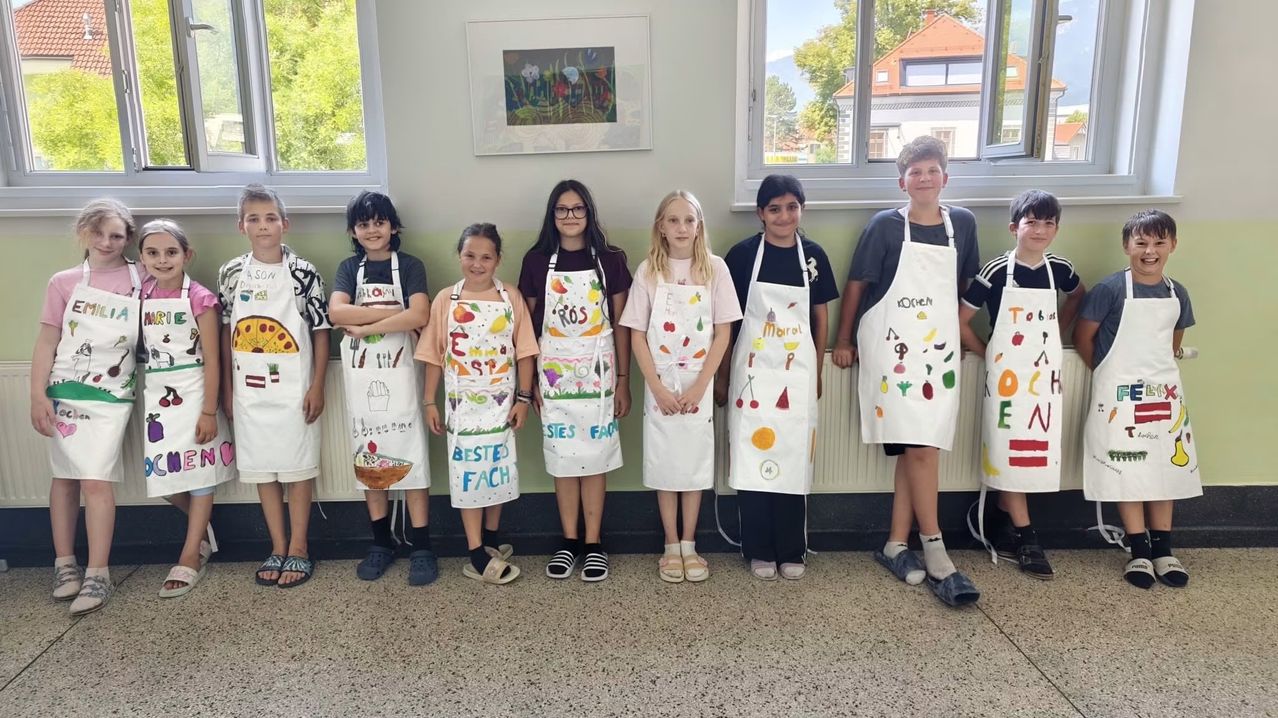 Eine Gruppe von Kindern in einem Klassenzimmer, die weiße Schürzen tragen, posiert für ein Foto. Jede Schürze hat bunte Zeichnungen und Texte. Dahinter ist eine Wand mit einem gerahmten Bild und Fenstern zu sehen.