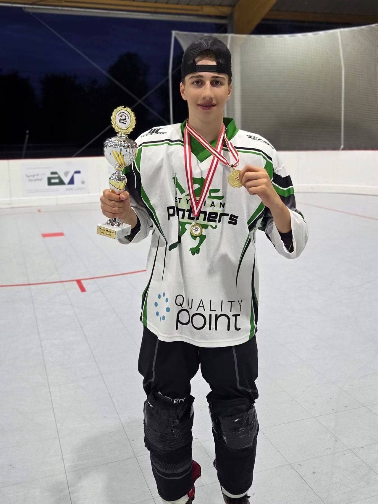 Ein junger Mann steht in einer Eishalle, hält einen Pokal und zwei Medaillen. Er trägt ein weißes und grünes Trikot mit 'QUALITY POINT' darauf. Er sieht glücklich und stolz aus.
