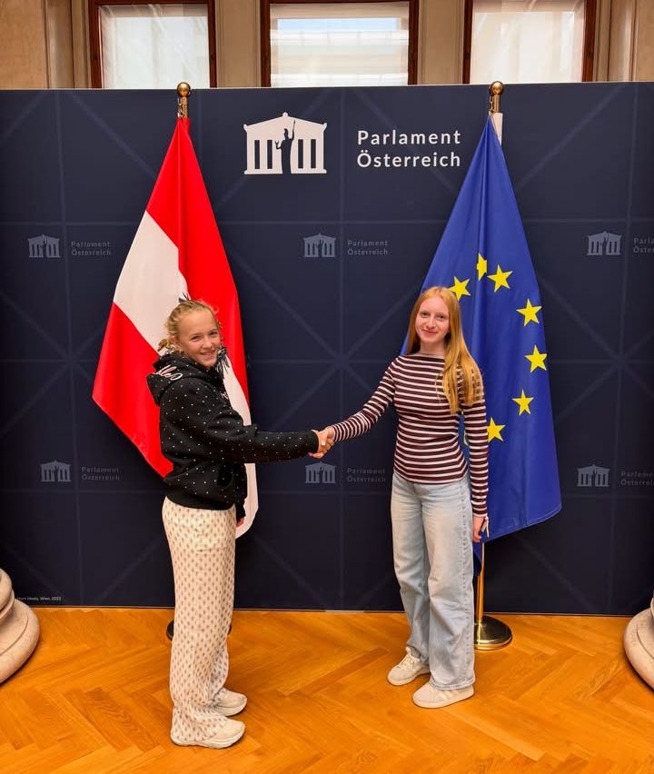 Zwei junge Frauen geben sich die Hand vor einem Hintergrund mit den Flaggen Österreichs und der Europäischen Union.