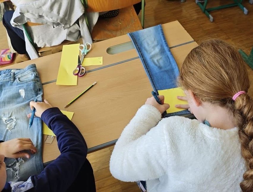Zwei Mädchen sitzen an einem Schreibtisch und arbeiten an einem Paar Jeans. Eine schneidet mit Schere und die andere markiert mit einem Bleistift. Der Schreibtisch ist mit Papieren und Stoff übersät.