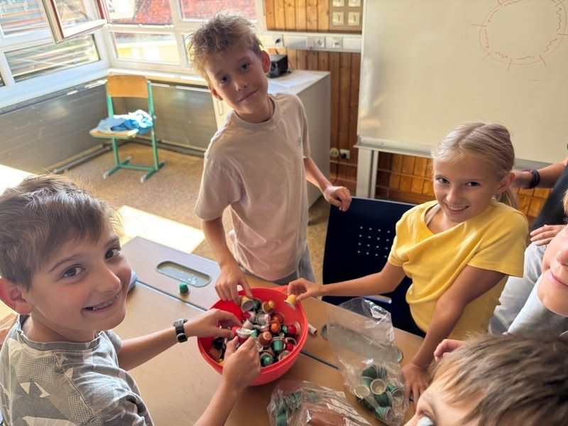 Eine Gruppe von Kindern versammelt sich um einen Tisch, lächelt und schaut in einen roten Eimer voller bunter Gegenstände.