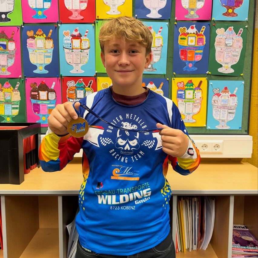 Ein junger Junge steht in einem Klassenzimmer und hält eine Medaille. Er trägt ein blau-weißes Radtrikot mit den Worten 'Wildfire' und 'Erdbau-Transporte' darauf. Hinter ihm befindet sich eine Wand mit bunten Eisbecher-Zeichnungen.