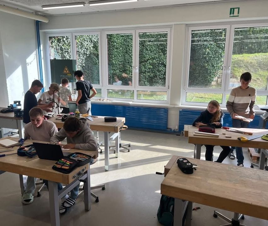 Eine Gruppe von Schülern arbeitet in einer Werkstatt an einem Projekt. Sie sitzen an Holztischen mit Laptops und verschiedenen Werkzeugen.