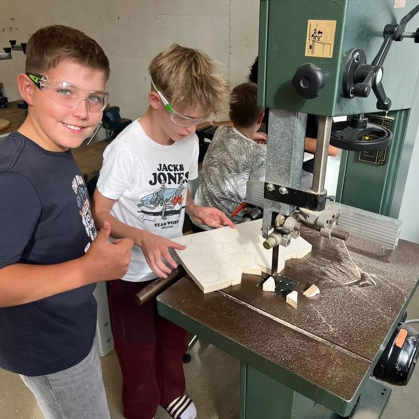 Zwei junge Jungen, die Schutzbrillen tragen, arbeiten in einer Werkstatt mit einer Bandsäge an einem Stück Holz.