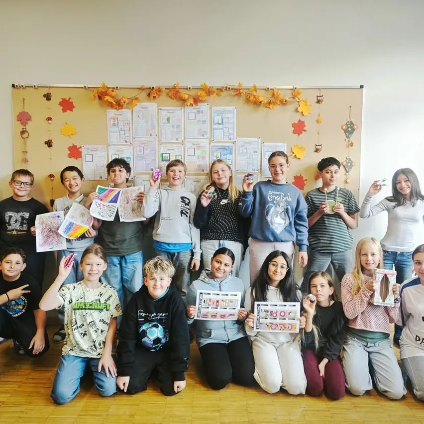 Eine Gruppe von Kindern posiert für ein Foto im Klassenzimmer. Sie halten Papierstücke mit Zeichnungen und Text hoch.