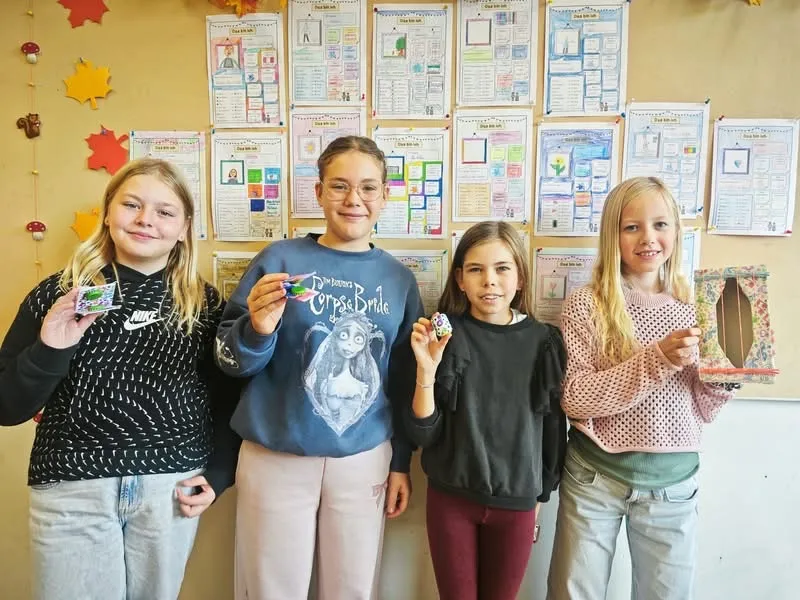 Vier Mädchen stehen vor einer Wand mit bunten Postern. Das Mädchen auf der linken Seite hält ein kleines Objekt, und das Mädchen in der Mitte hält ein kleines Objekt mit einem Bild eines Mädchens darauf.