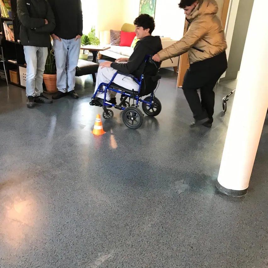 Eine Gruppe von Menschen versammelt sich in einem Raum mit einer Person im Rollstuhl. Die Person im Rollstuhl wird von einer anderen Person unterstützt. Es gibt Topfpflanzen und einen Bücherschrank im Raum.