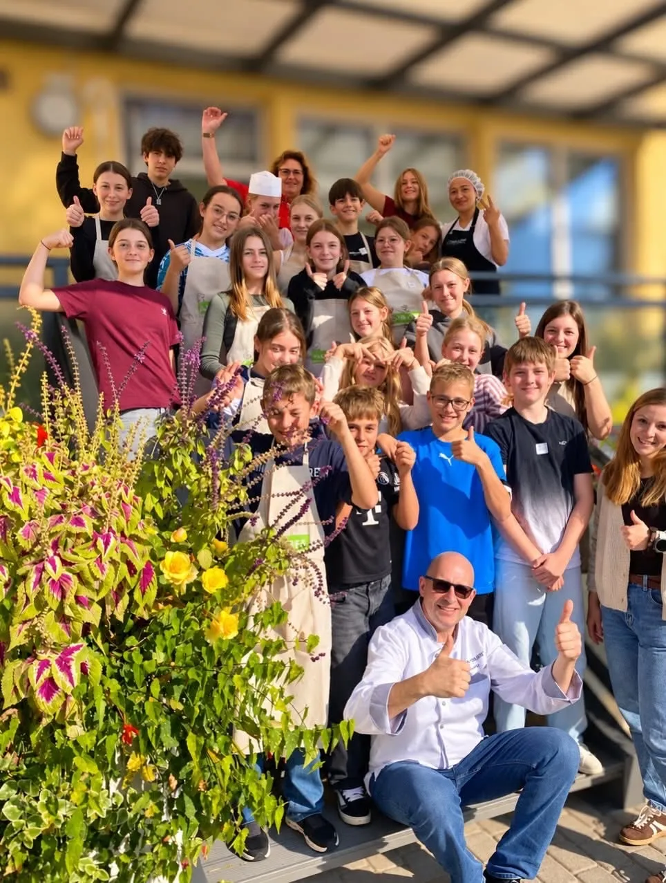 Eine Gruppe junger Menschen und Erwachsene posiert für ein Foto vor einem gelben Gebäude. Sie tragen alle Schürzen und einige zeigen Daumen hoch.