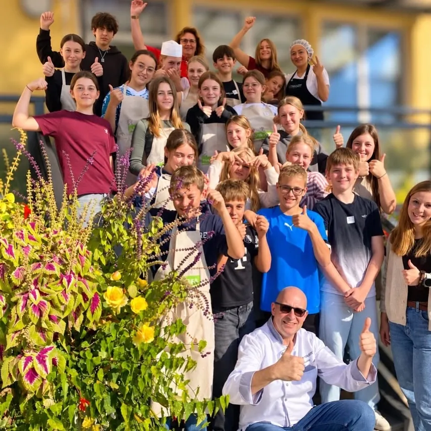 Eine Gruppe junger Menschen und Erwachsene posiert für ein Foto vor einem gelben Gebäude. Sie tragen alle Schürzen und einige zeigen Daumen hoch.