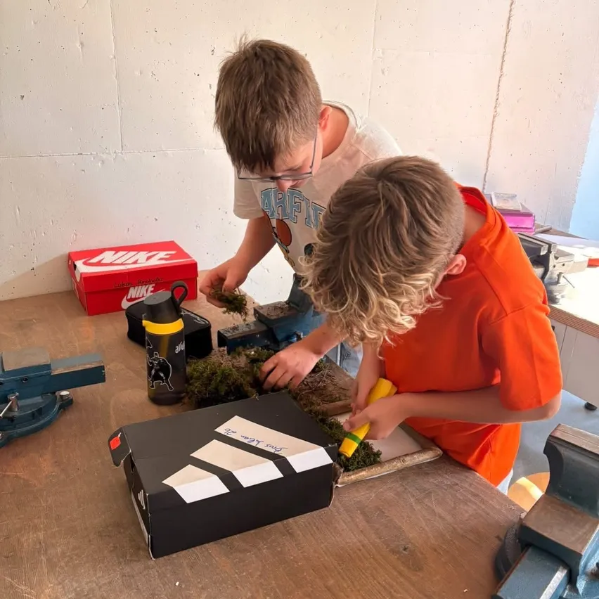 Zwei Jungen arbeiten in einer Werkstatt an einem Projekt. Einer trägt eine Brille und der andere ein orangefarbenes Shirt.