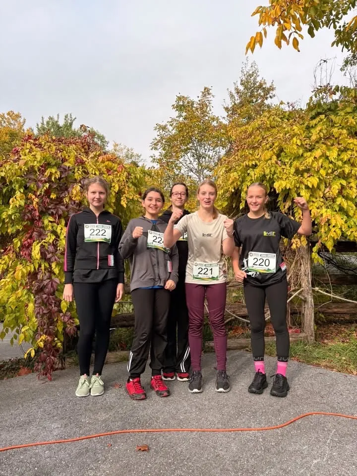 Fünf Frauen in Laufausrüstung stehen in einer Reihe, lächeln und posieren für ein Foto vor einer Reihe von Bäumen.