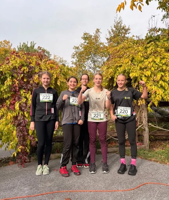 Fünf Frauen in Laufausrüstung stehen in einer Reihe, lächeln und posieren für ein Foto vor einer Reihe von Bäumen.