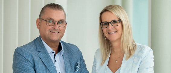 Ein Mann und eine Frau stehen gemeinsam, lächeln und posieren für ein Foto. Sie sind professionell gekleidet, und die Frau hat blonde Haare.